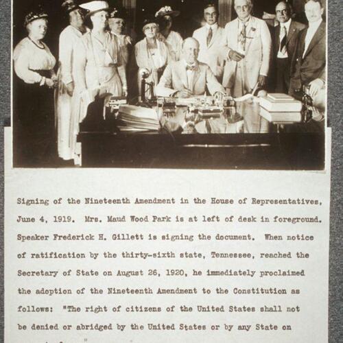 Gillett at desk, seated, poised as if signing a document, looking up toward camera. A group of women stand behind him to the left, a group of men stand behind him to the right.