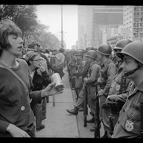 A young &quot;hippie&quot; standing in front of a row of National Guard soldiers, across the street from the Hilton Hotel at Grant Park, at the Democratic National Convention in Chicago